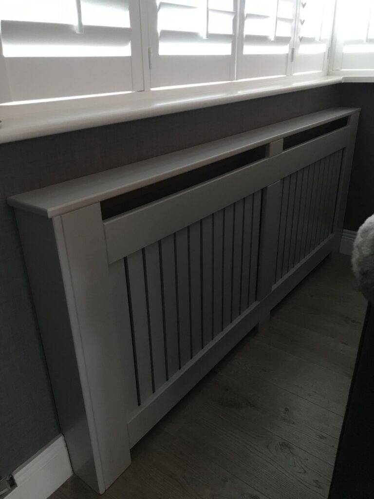 A light gray, modern radiator cover with vertical slats and a flat top shelf, positioned beneath a window with white plantation shutters.
