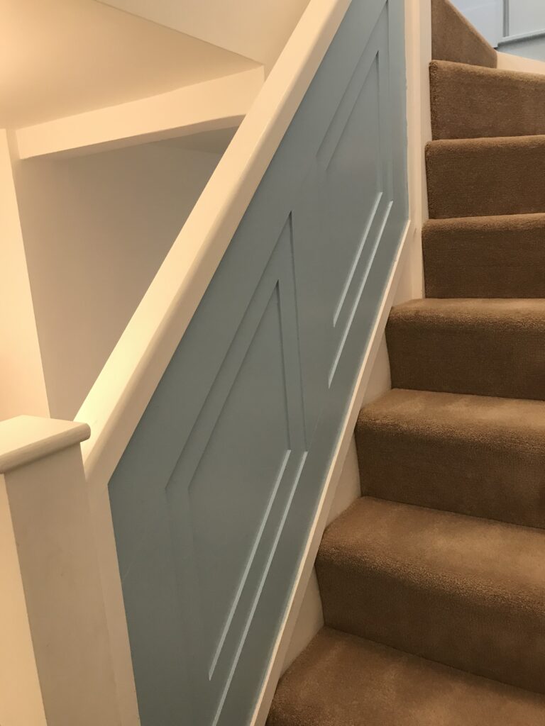 A staircase with grey-carpeted steps. The solid wall-like banister is painted a soft baby blue and features a decorative geometric panel pattern created with raised molding.